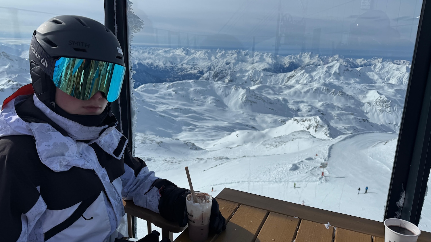 Daniel drinking a hot chocolate in full ski attire with a snow covered mountain backdrop