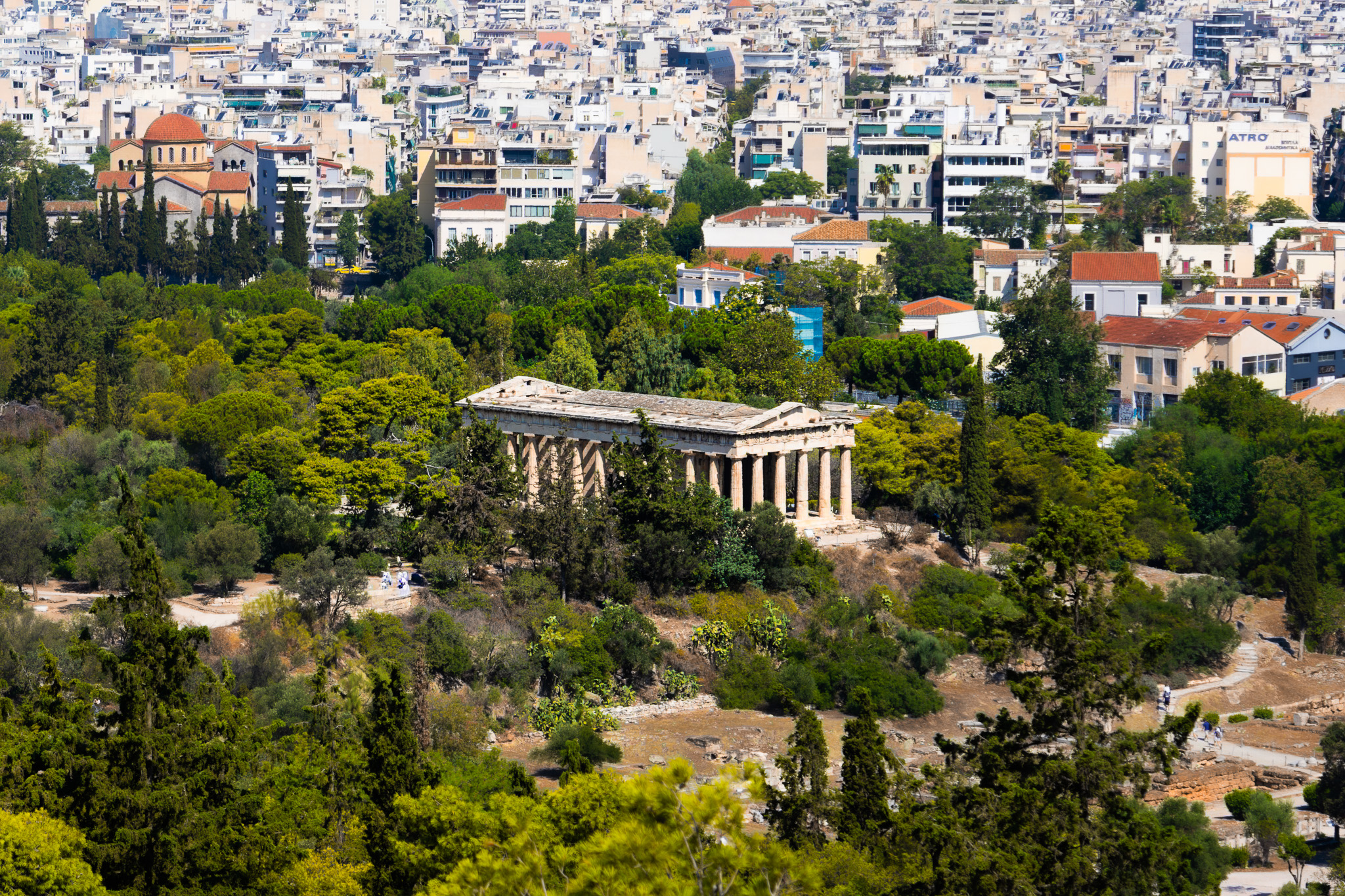 An ancient greek temple surrounded by trees bordered by a bustling city